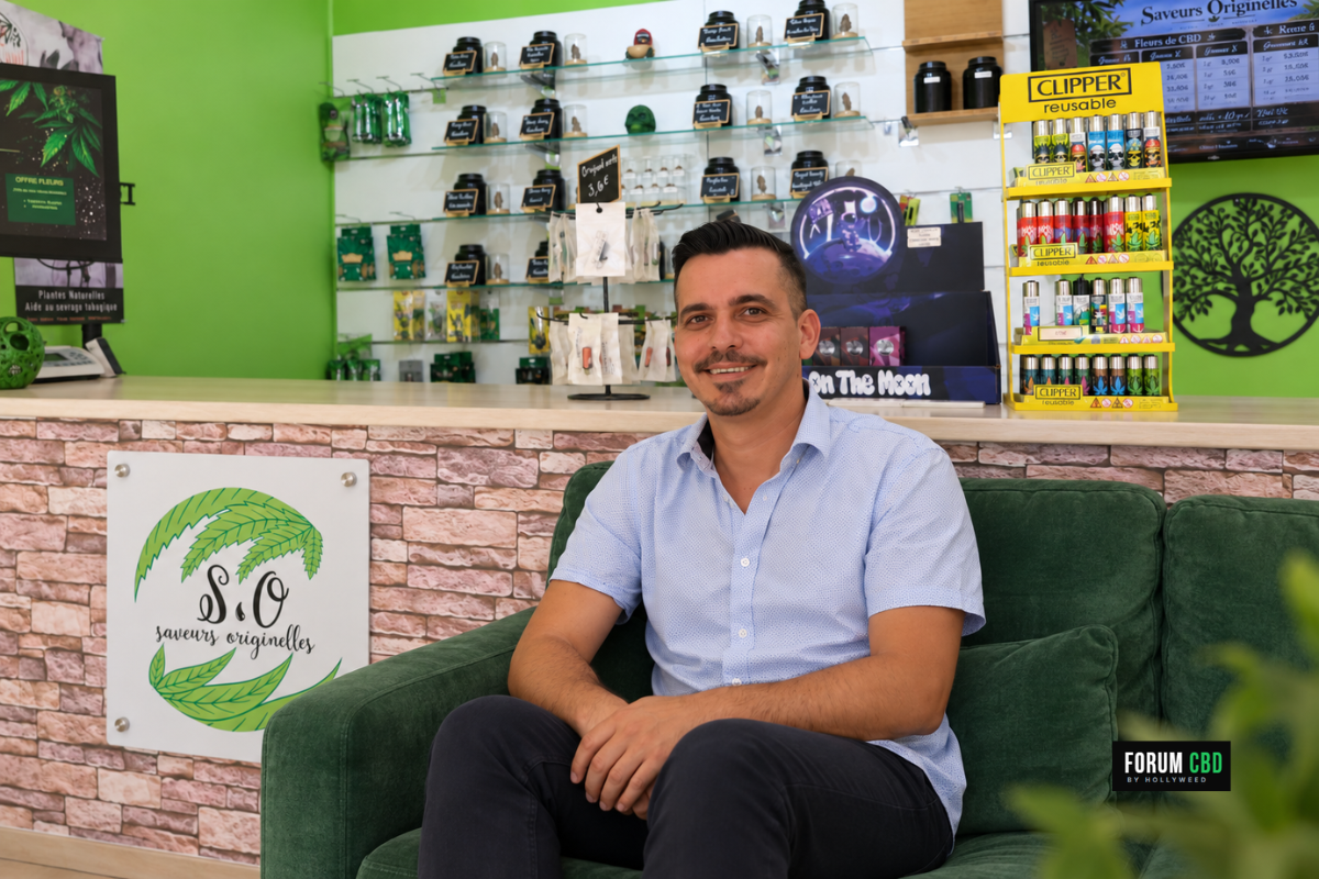 Mise en scène de Jimmy Boada assis dans un sofa vert devant le comptoir et le logo de sa boutique Saveurs Originelles à Orléans.