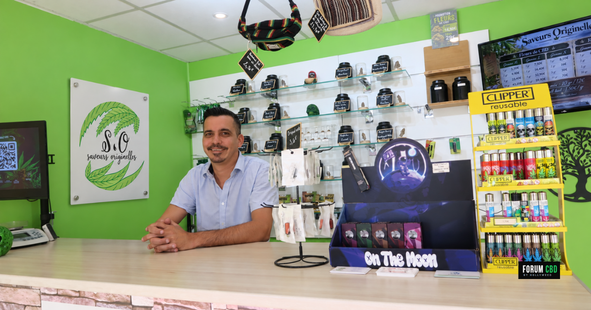 Commerçant derrière le comptoir de la boutique Saveurs Originelles, avec les produits en présentation et une plaque du logo sur le mur vert.